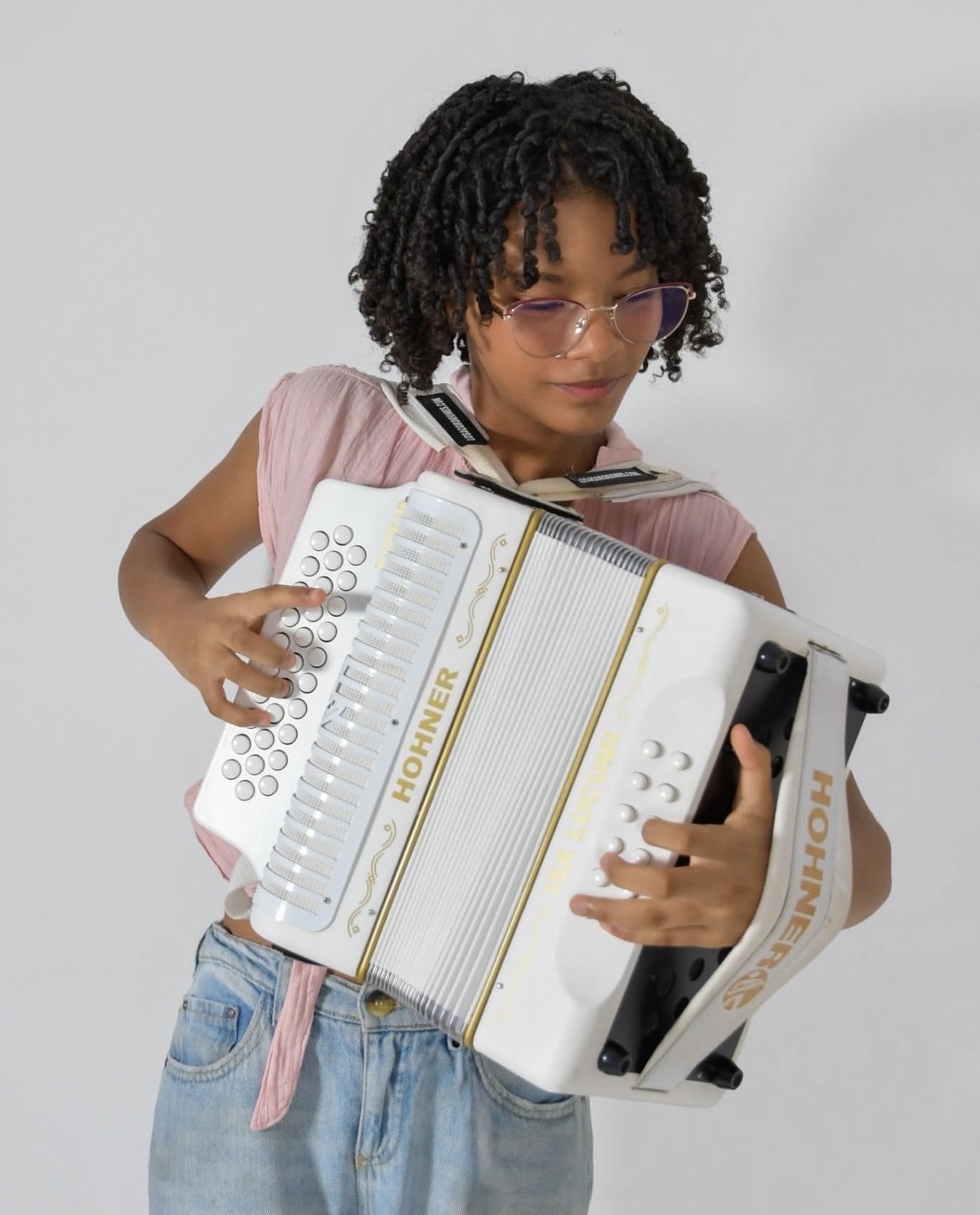 Niña con cabello rizado y gafas tocando un acordeón blanco frente a un fondo claro.
