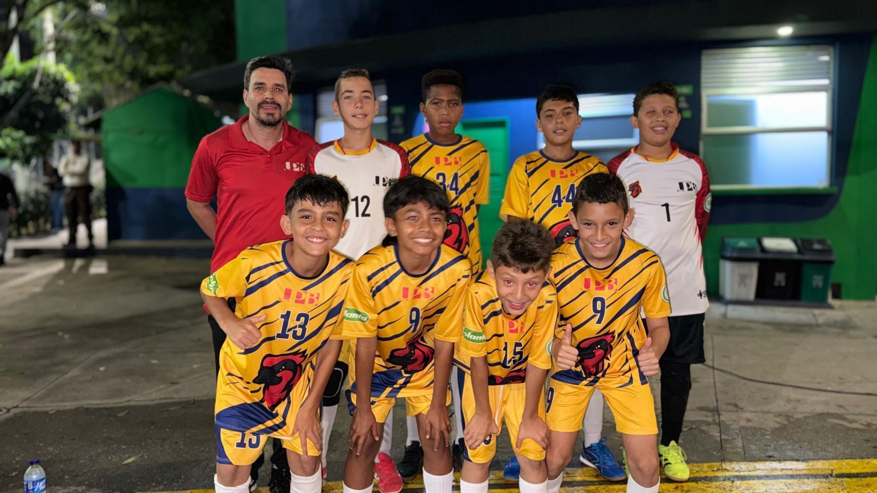 Un equipo infantil de fútbol posa sonriente junto a su entrenador por la noche, vistiendo uniformes amarillos con azul de la UPB y uniformes blancos de portero.
