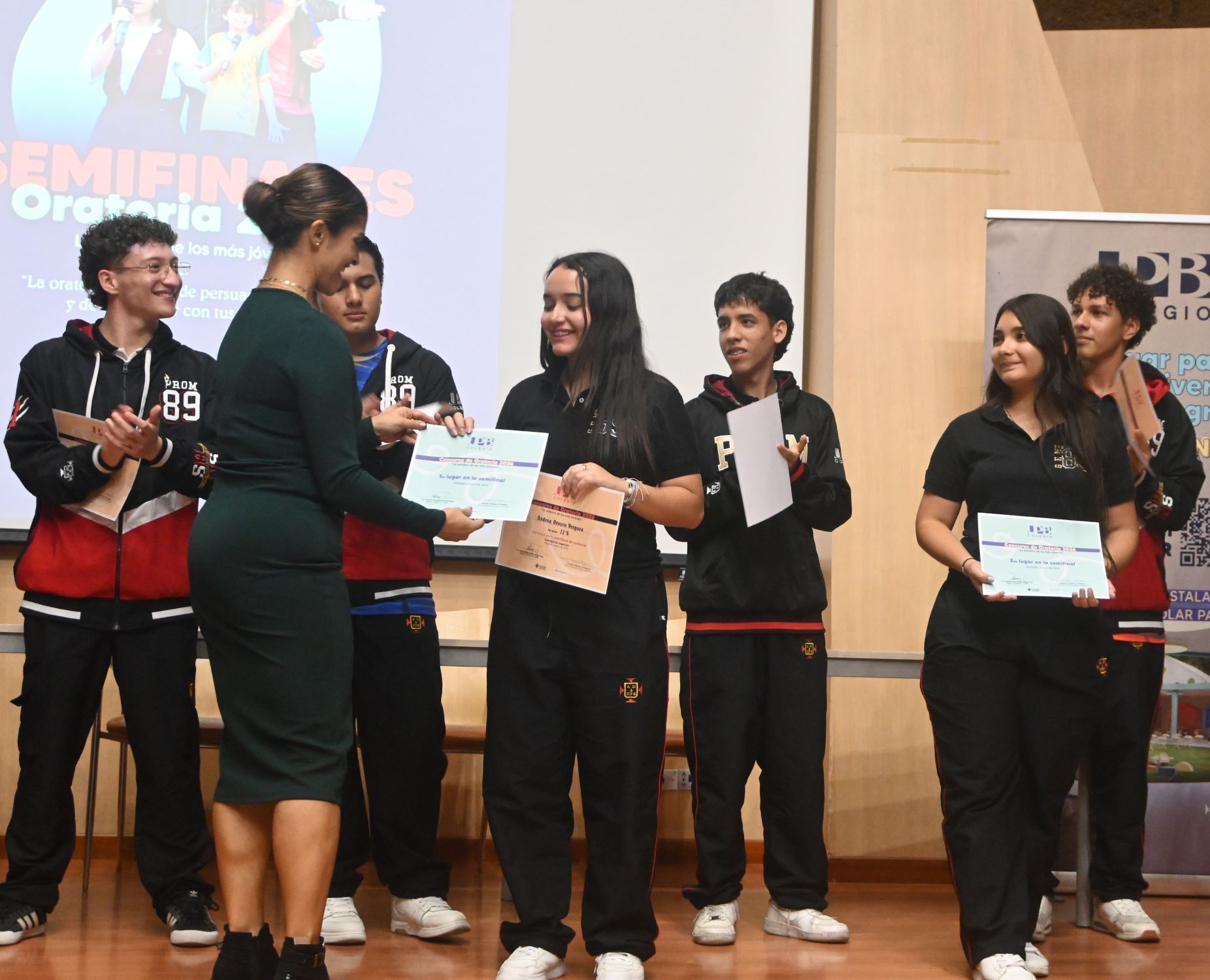 Estudiantes uniformados reciben certificados de oratoria en un escenario durante una ceremonia escolar.