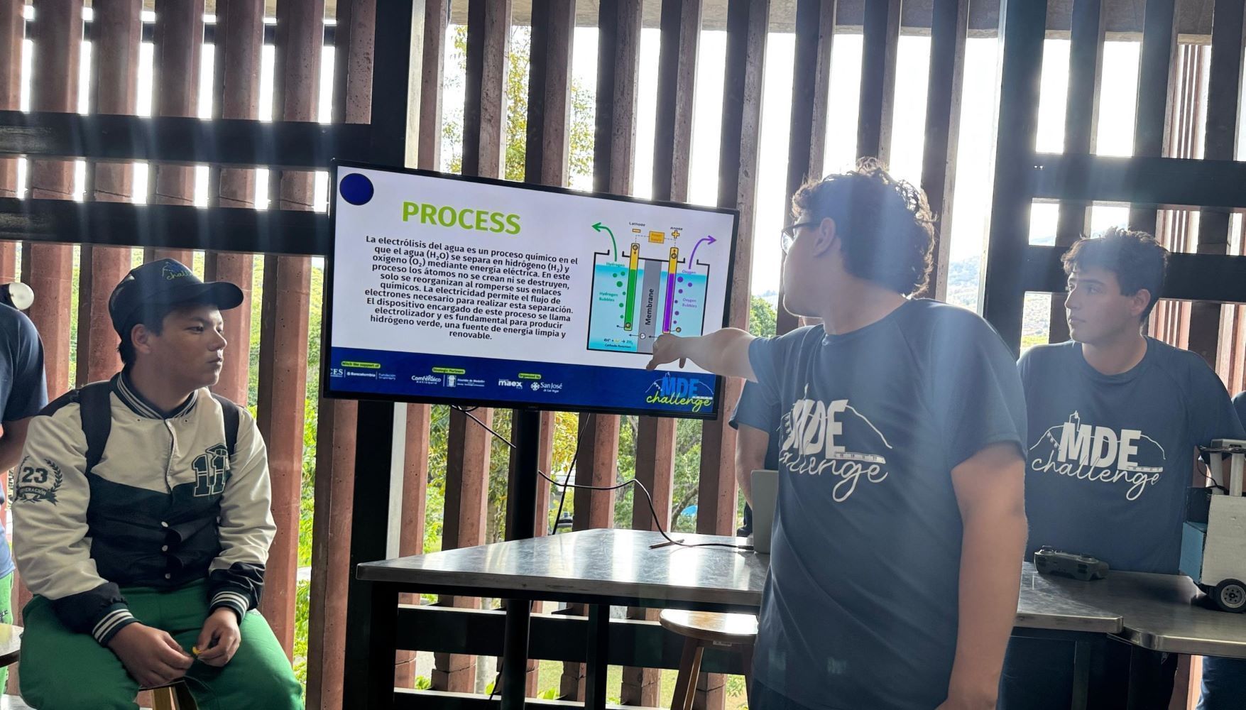 Un estudiante expone sobre el proceso de electrólisis del agua señalando un diagrama explicativo en una pantalla, mientras otros jóvenes escuchan.