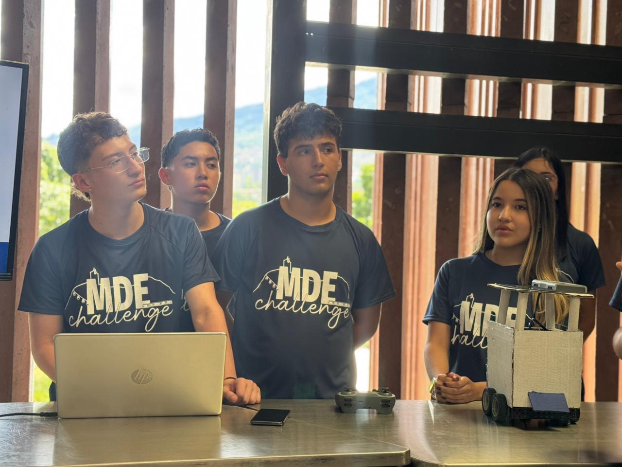 Cinco jóvenes con camisetas del "MDE challenge" atienden a una explicación en un taller, junto a una laptop, un control remoto y un prototipo móvil sobre la mesa.