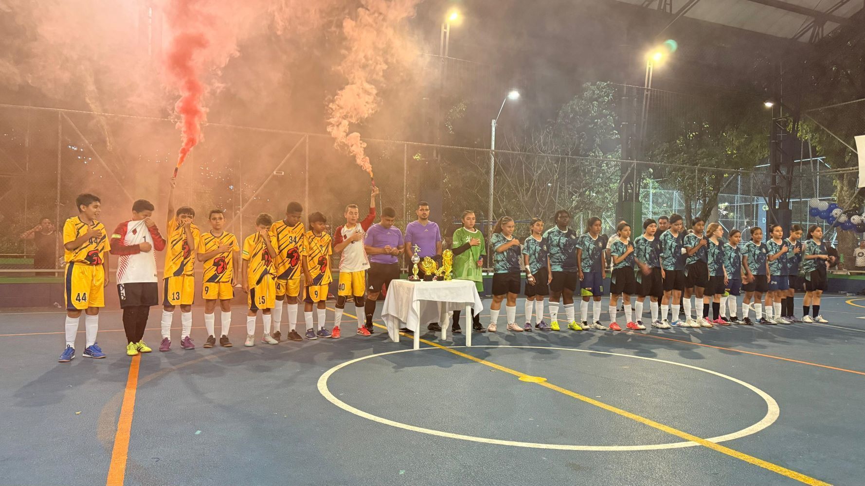 Dos equipos juveniles de fútbol sala alineados en la cancha durante una ceremonia de premiación nocturna, con trofeos en una mesa y bengalas de humo rojo de fondo.