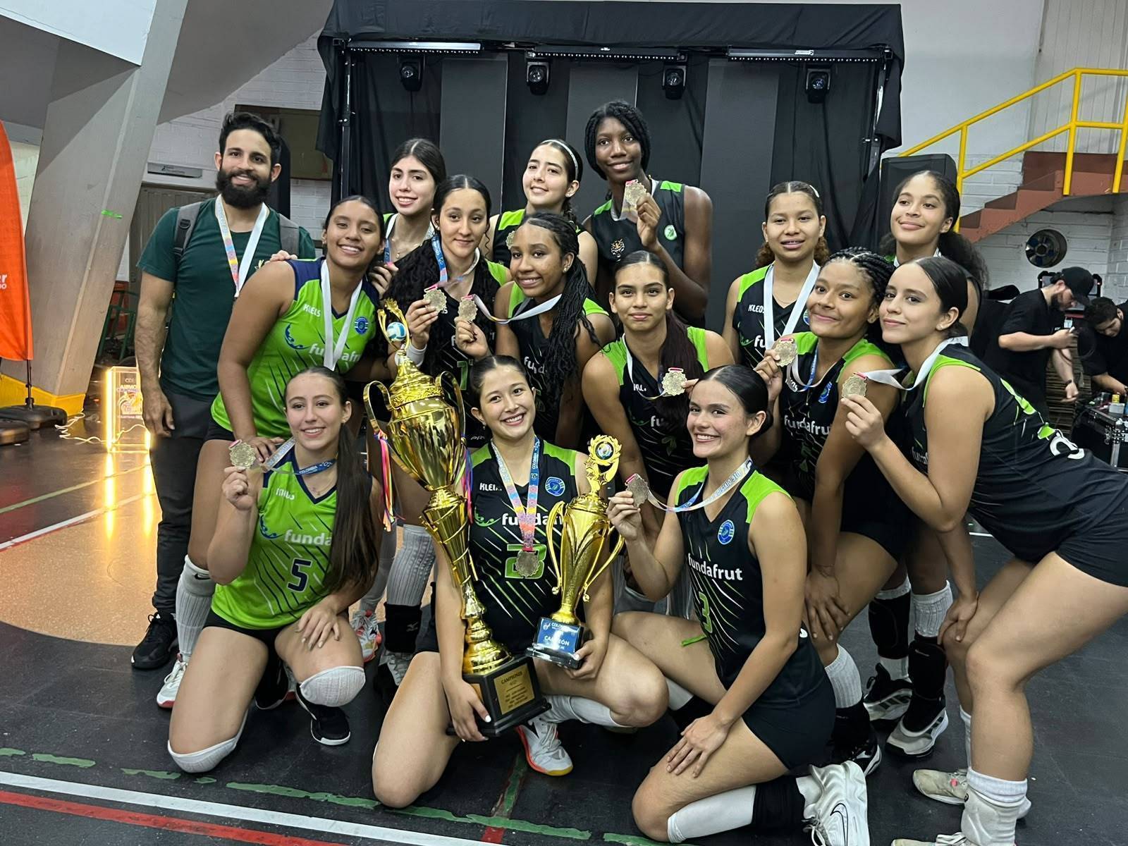 Equipo de voleibol femenino de Fundafrut posa sonriente con trofeos y medallas de campeonas en una cancha cubierta.