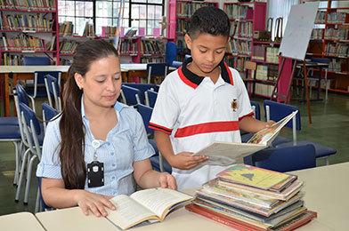 Una asistente de la Biblioteca está sentada leyendo un libro y a su lado se encuentra un estudiante de pie leyendo otro. Frente de ellos se encuentra una pila de libros sobre una mesa.