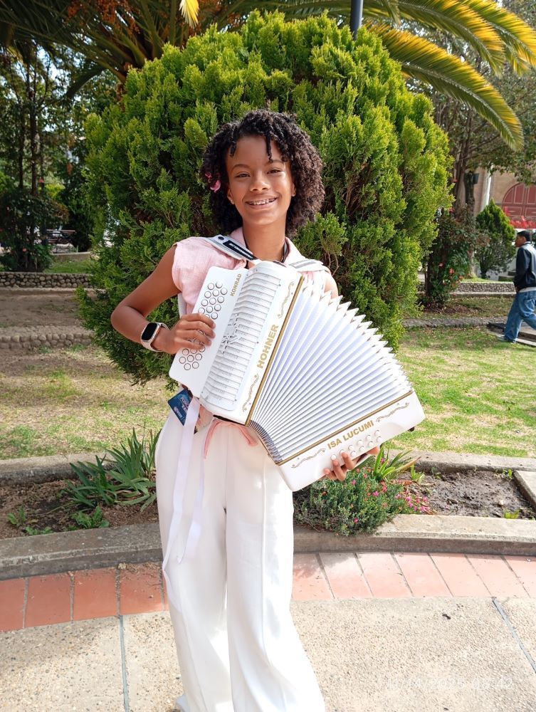 Una niña alegre vestida de blanco y rosa sostiene un acordeón blanco abierto frente a un arbusto verde en un parque.