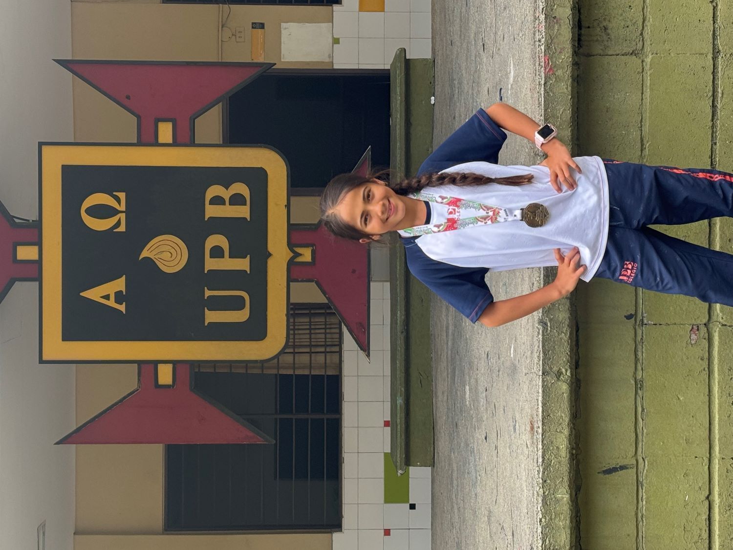 Niña con uniforme escolar y medalla, sonriendo frente al escudo del colegio UPB.
