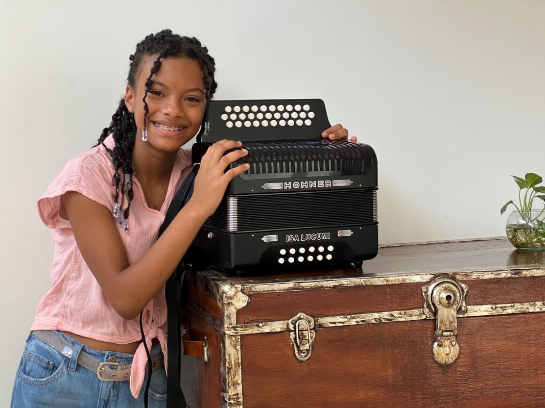 Una niña sonriente con trenzas posa junto a un acordeón negro, el cual está apoyado sobre un baúl de madera antiguo.