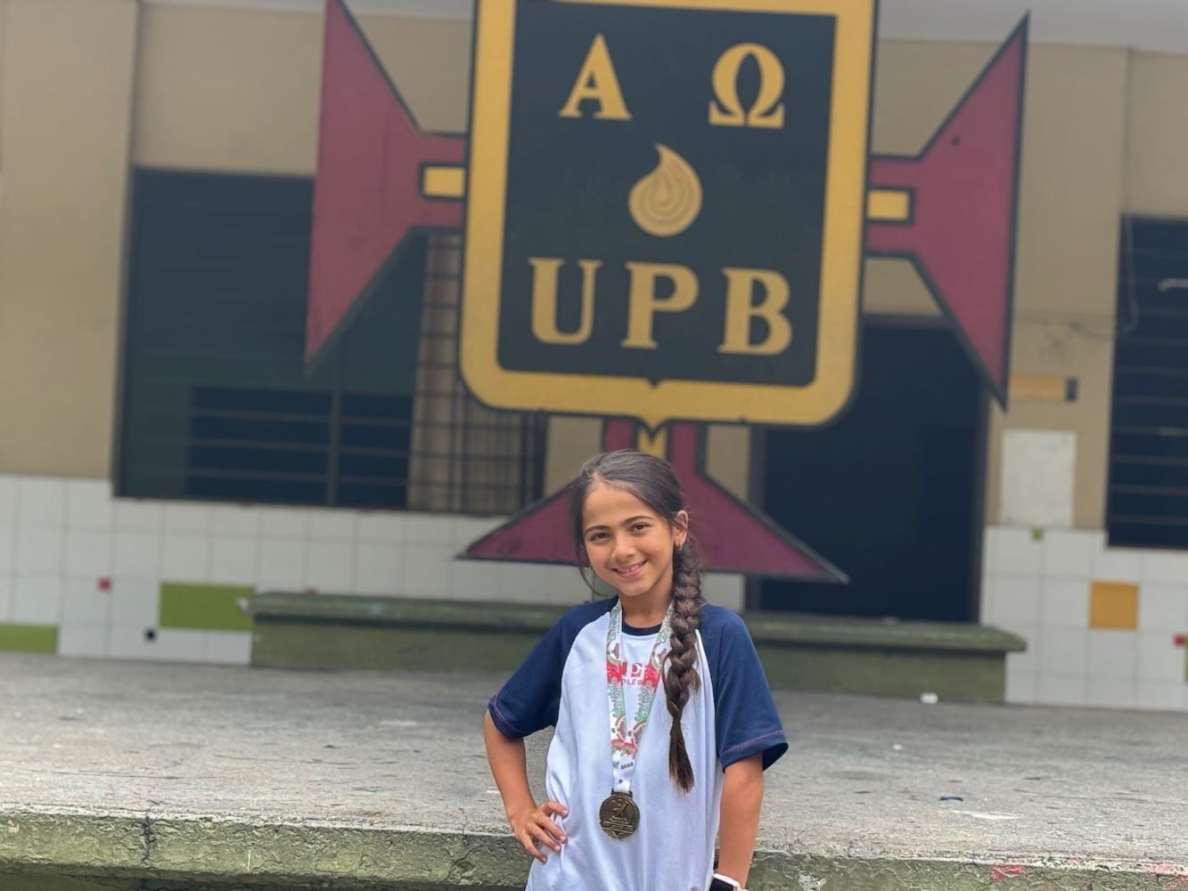 Niña con uniforme escolar y medalla, sonriendo frente al escudo del colegio UPB en un patio.