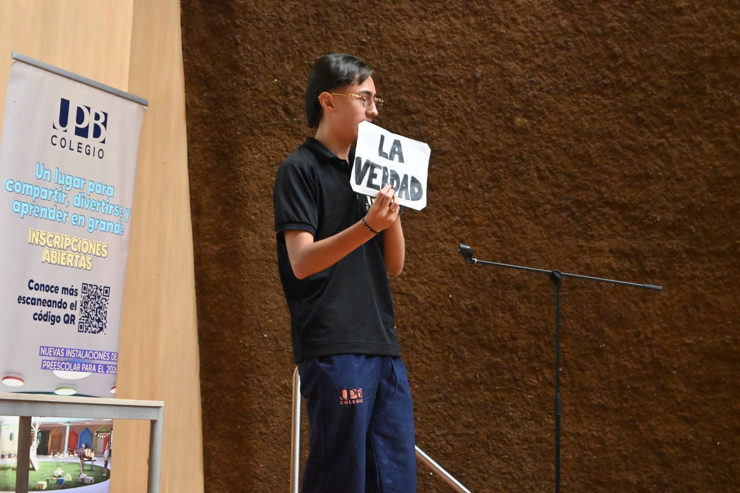 Estudiante del Colegio UPB de pie en un auditorio, sosteniendo un cartel con el texto "LA VERDAD" cerca de su rostro durante una presentación.
