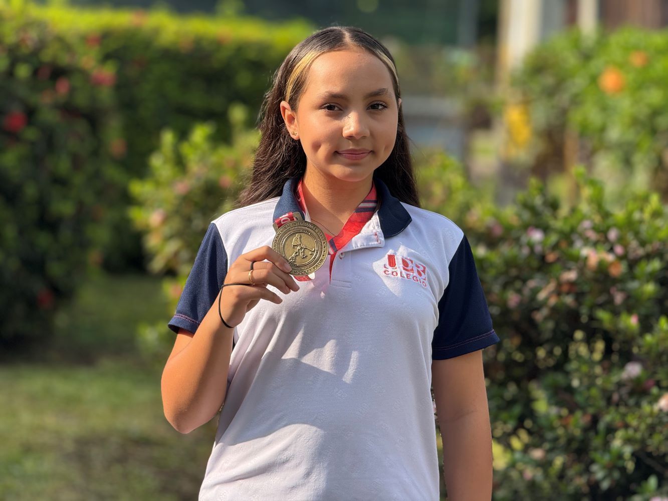 Estudiante del Colegio UPB posando con su medalla deportiva en un jardín.