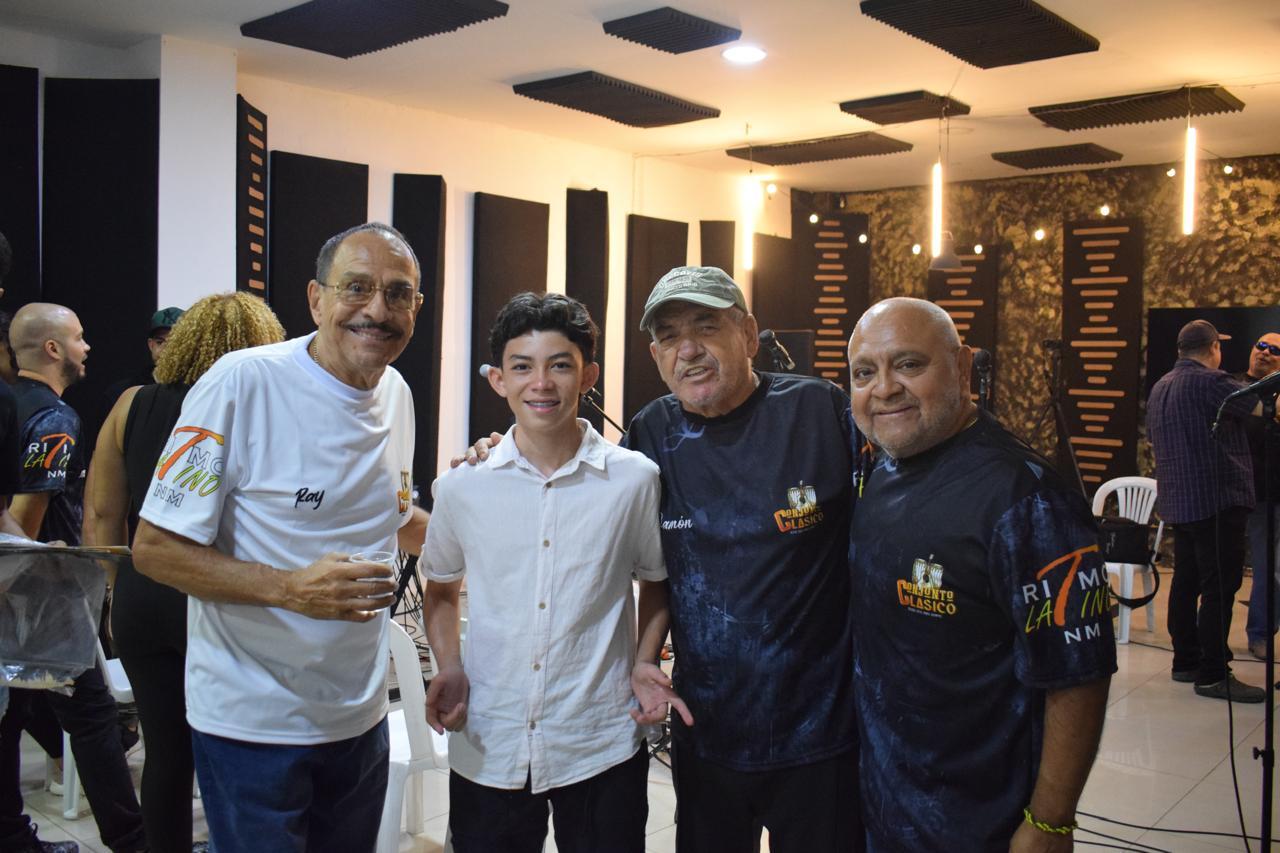 Un joven con camisa blanca posa sonriente junto a tres hombres adultos en un estudio de grabación musical.