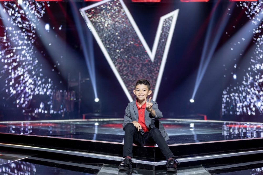 Un niño sentado sonriendo en el escenario de La Voz, haciendo la señal de la victoria con los dedos frente al gran logo de la "V".