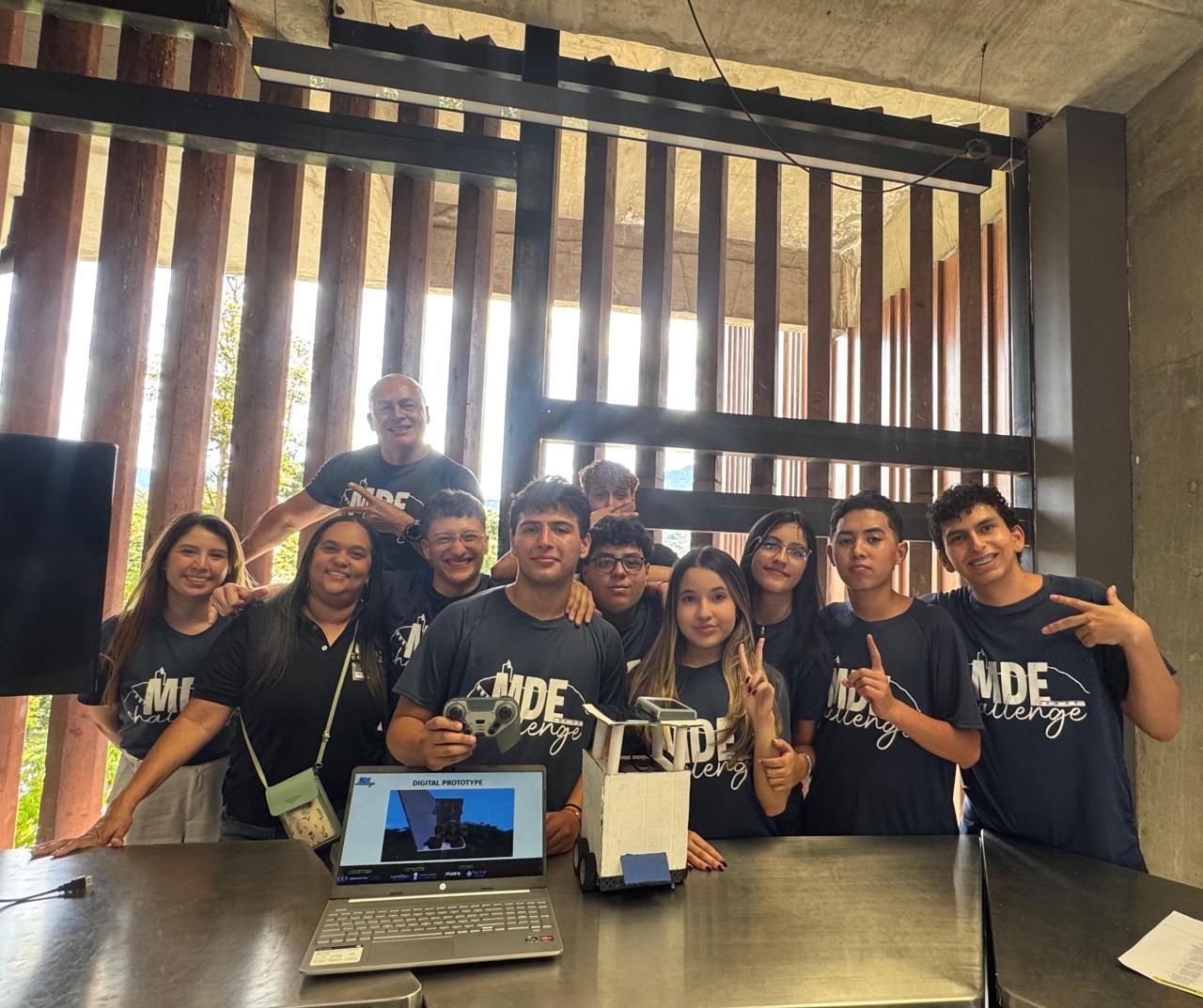 Grupo de jóvenes y mentores sonriendo junto a una laptop y un prototipo tecnológico en un taller con fondo de vigas de madera.