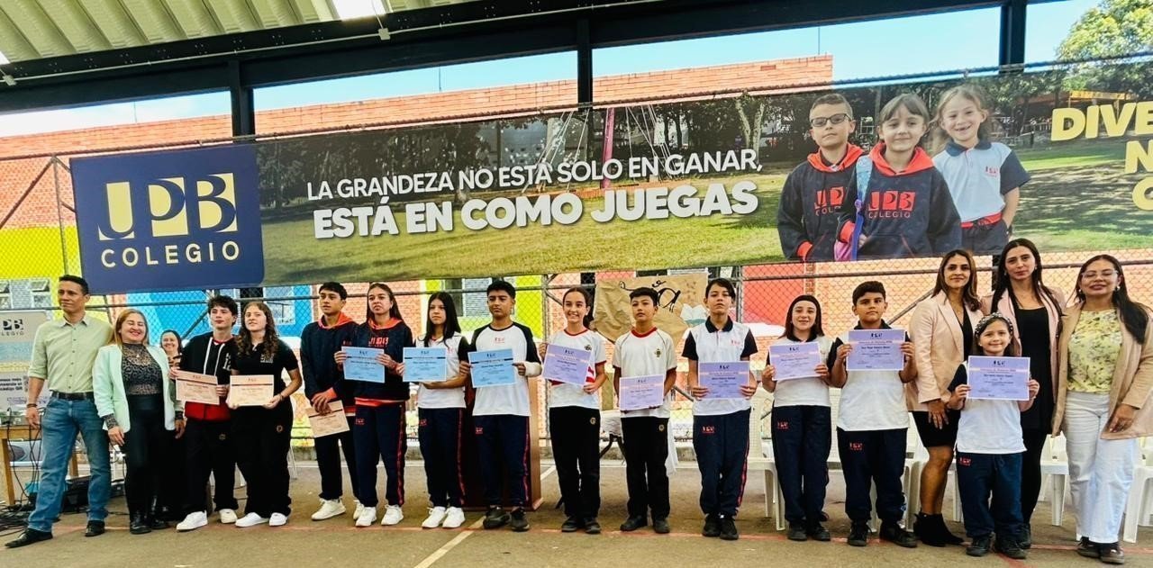 Alumnos y profesores del Colegio UPB posan alineados en una cancha abierta mostrando certificados, con una lona publicitaria de fondo.