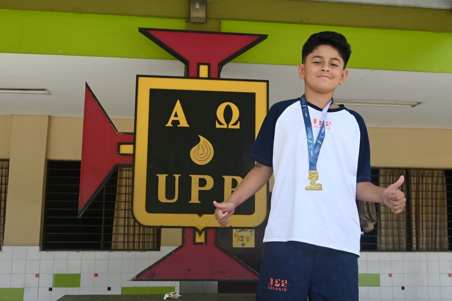 Un estudiante con uniforme del Colegio UPB posa con una medalla de oro y los pulgares arriba frente al gran escudo de la institución.