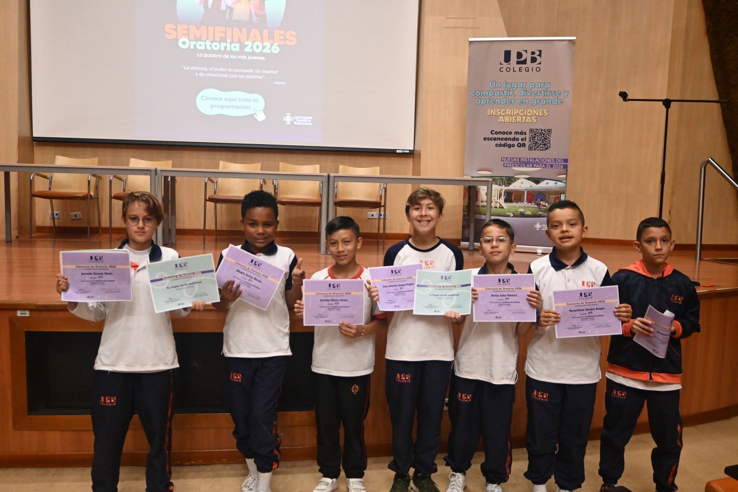 Un grupo de siete estudiantes del Colegio UPB posa frente al escenario de un auditorio sosteniendo certificados del concurso "Semifinales Oratoria 2026"