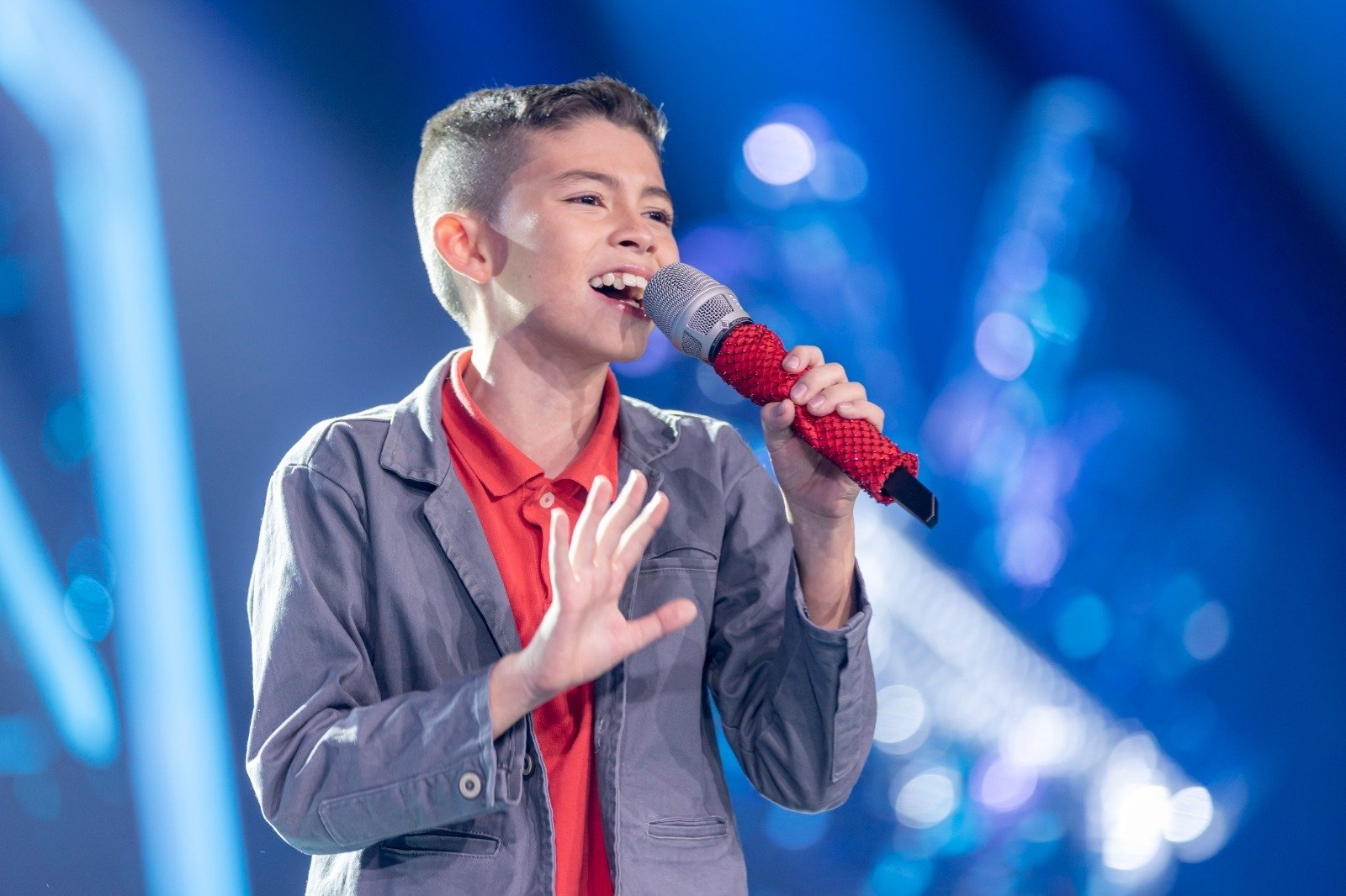 Un niño cantando apasionadamente con un micrófono rojo en un escenario con luces azules de fondo.