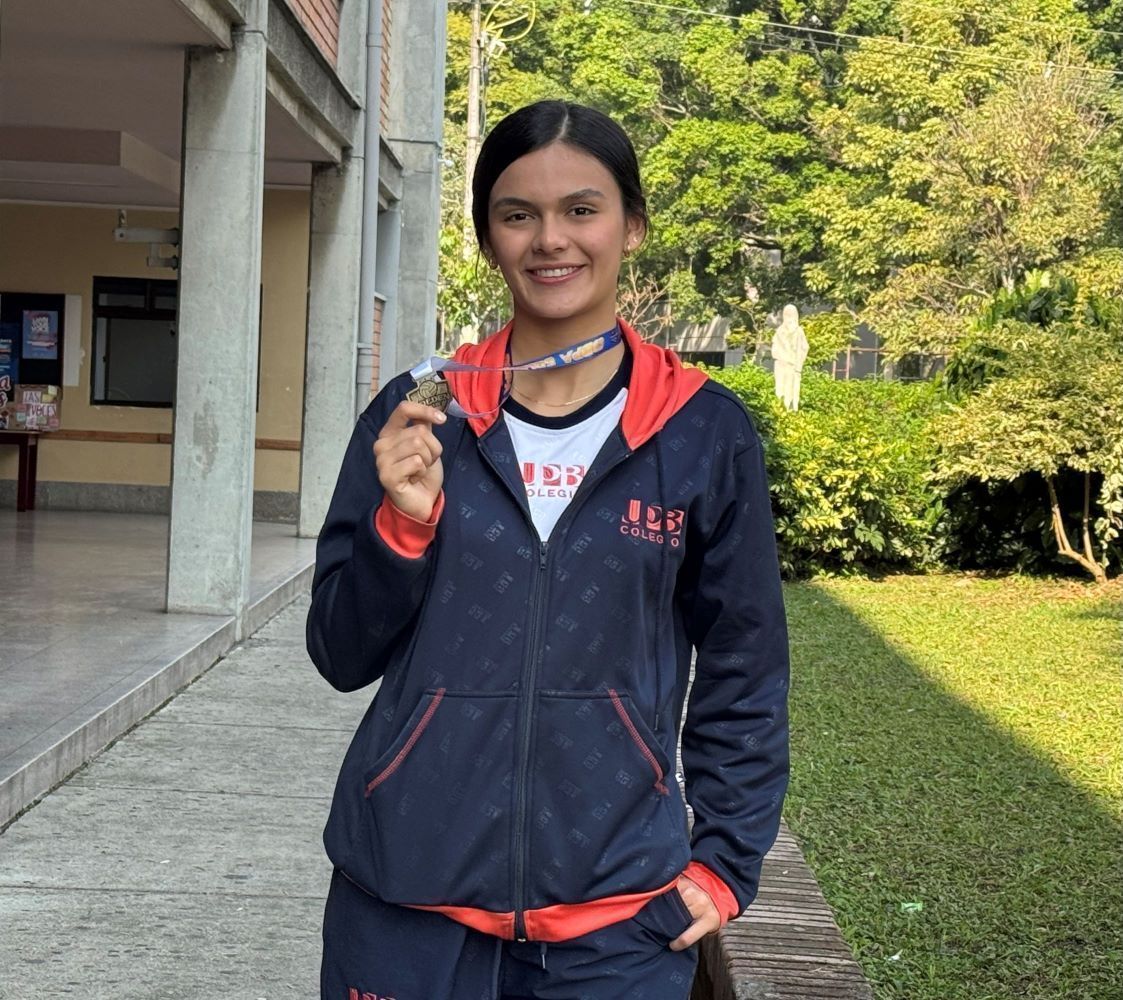 Una joven con uniforme del Colegio UPB sonríe orgullosa sosteniendo una medalla plateada que cuelga de su cuello. Posa de pie en un pasillo exterior del colegio, con zonas verdes y una estatua religiosa al fondo.