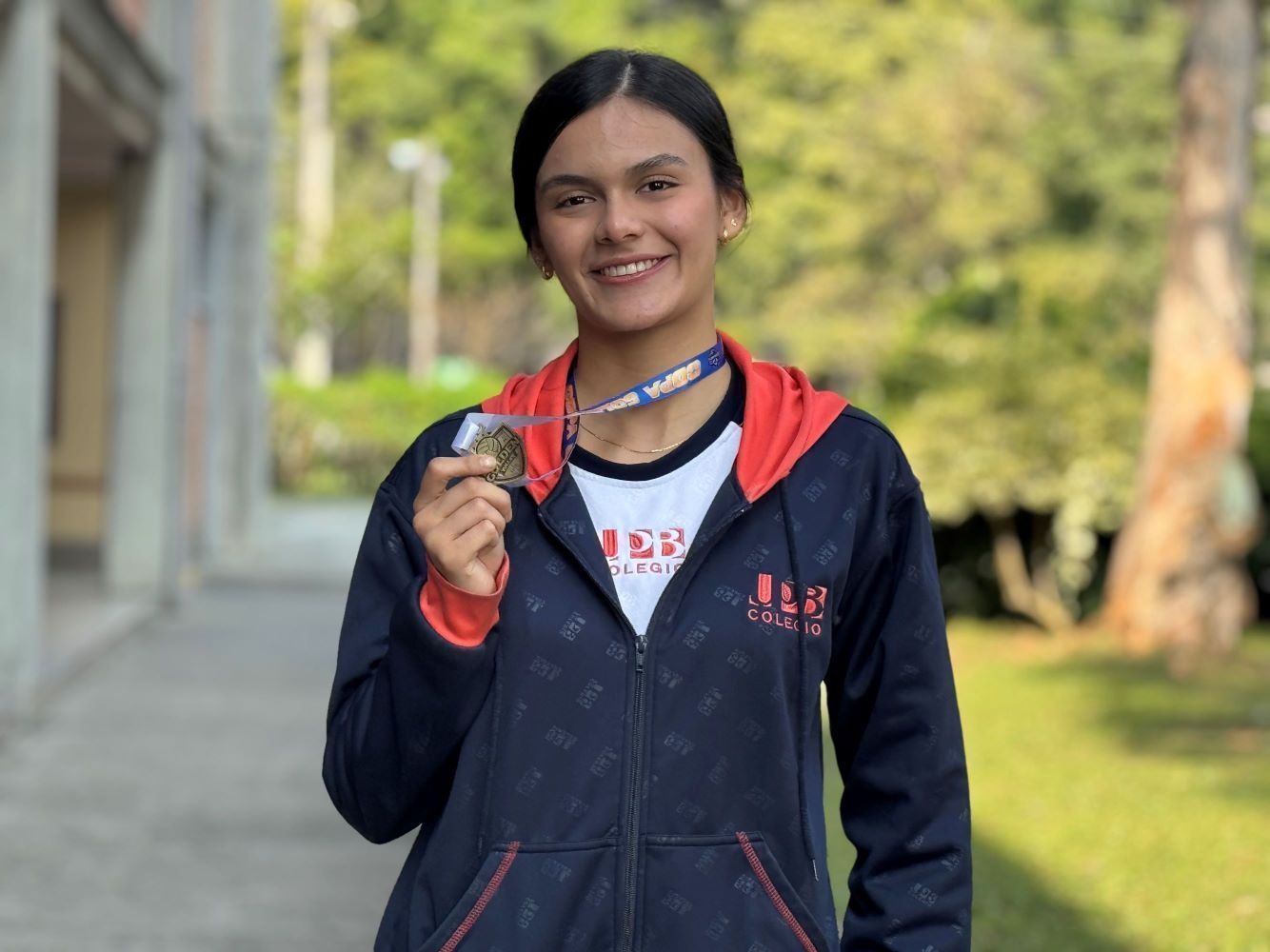 Una joven con el uniforme del Colegio UPB sonríe mientras sostiene una medalla dorada, posando en un entorno escolar al aire libre.