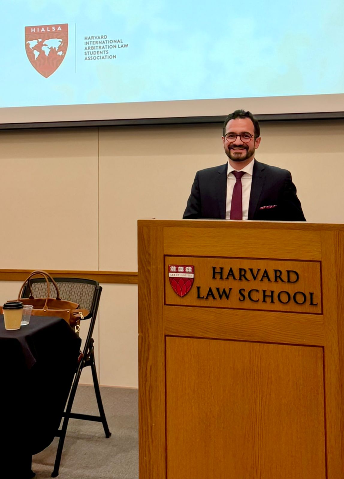 Alirio Giraldo sonriendo tras un podio con el logo de Harvard Law School durante una conferencia de arbitraje internacional.