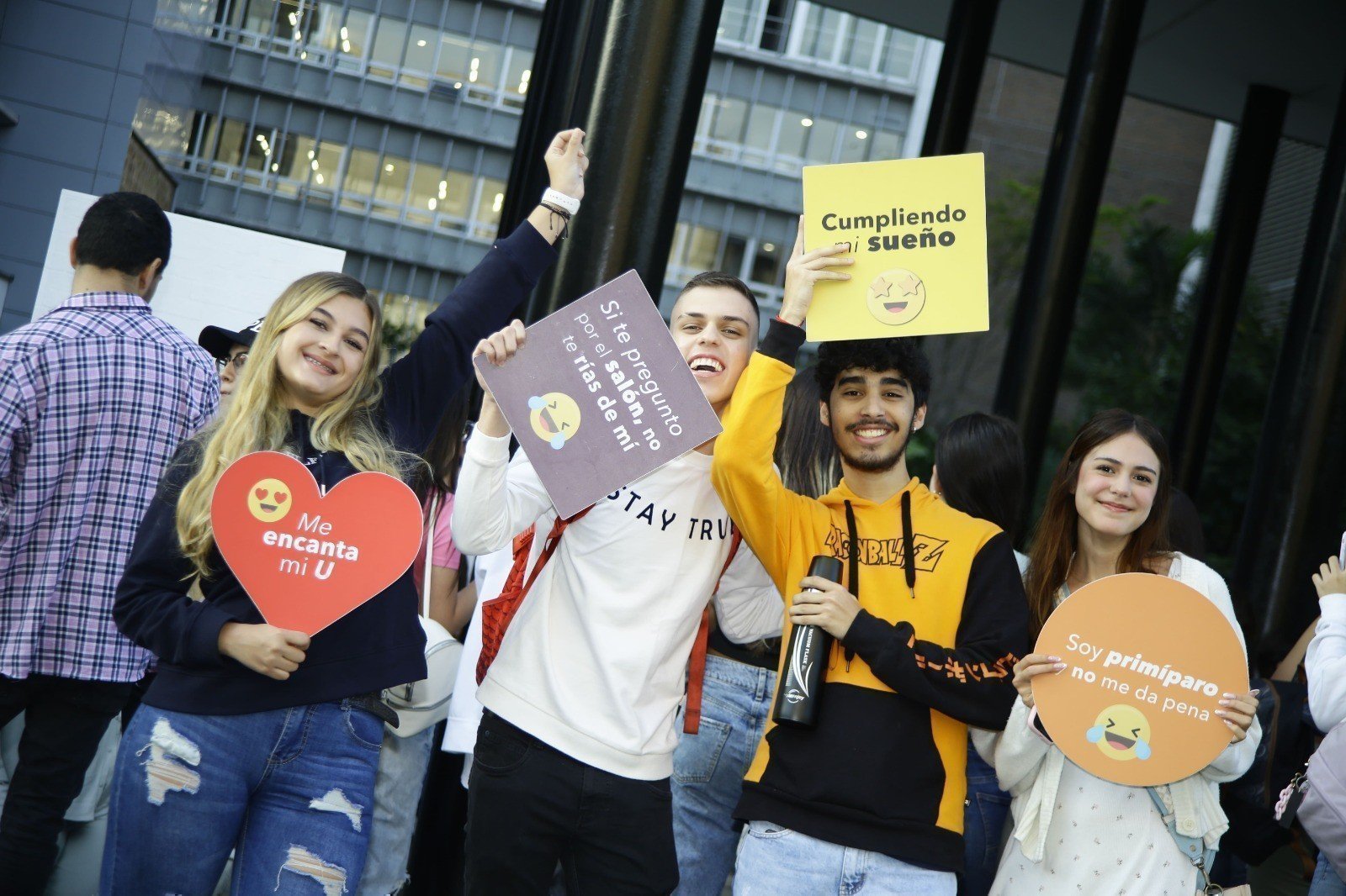 Cuatro estudiantes alegres sosteniendo carteles de emoticones con mensajes de bienvenida a la UPB
