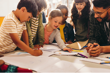 Un docente con gafas guía a un grupo diverso de niños que trabajan con papeles y cuadernos alrededor de una mesa, en un espacio luminoso de aprendizaje colaborativo.
