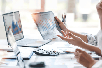 Dos personas analizan datos en una mesa: una sostiene una tablet con un gráfico de barras titulado "Sale performance" mientras la otra gesticula durante la conversación, con un portátil, una calculadora y documentos con gráficos bajo la luz del sol.