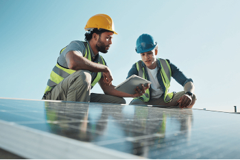 Dos trabajadores de la construcción con cascos y chalecos de seguridad revisan una tablet en una azotea, en primer plano unos paneles solares oscuros y un cielo azul despejado.