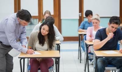 docente se acerca a una alumna y le explica algo. El resto de estudiantes escriben