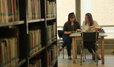 estantería con libros. Al fondo se ven dos estudiantes estudiando en una mesa