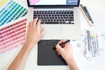Mano sosteniendo lapiz sobre computador, al lado impresiones de tabletas de colores