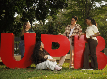 Cuatro jóvenes posan sonrientes al aire libre junto a las grandes letras rojas "UPB" sobre el césped, con árboles al fondo.