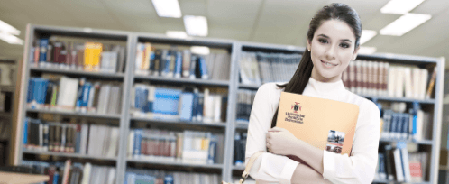 Mujer sosteniendo una carpeta con diseño UPB, al fondo se ve una biblioteca con libros