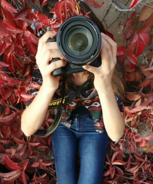 Persona acostada sobre unas hojas rojas sostiene una camara de fotografía como si tomara una foto hacia el cielo