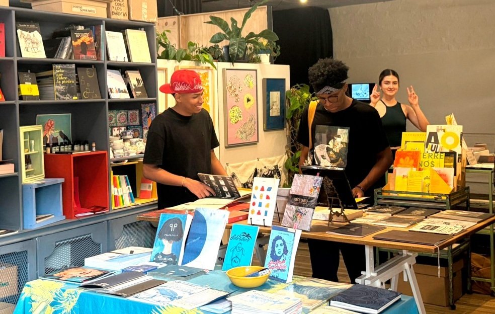 Mesa de venta de libros y arte con personas observando y sonriendo; fondo de estanterías y vitrinas con publicaciones coloridas.