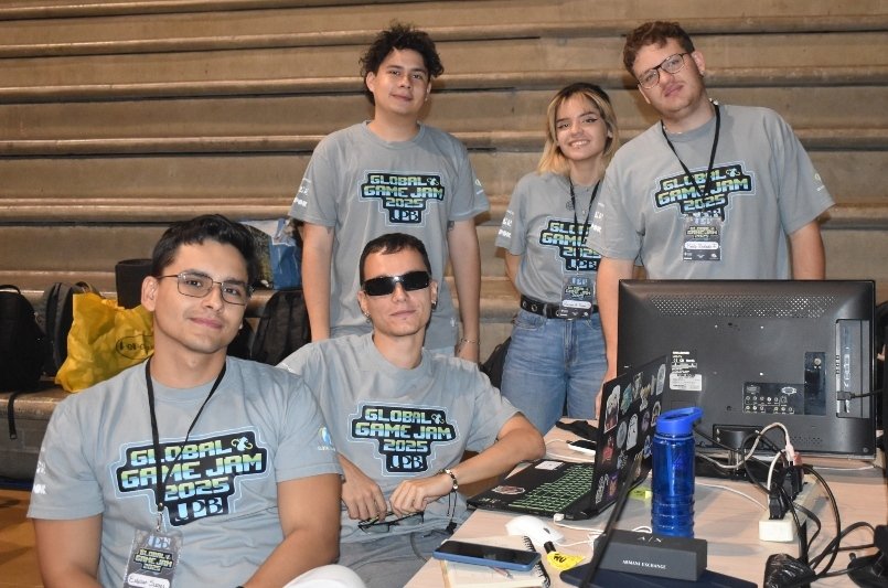 Grupo de jóvenes en un puesto de videojuegos, con camisetas grises y lanyards, trabajando frente a computadoras y monitor en un evento.