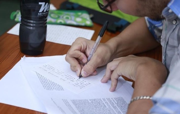 Persona escribiendo notas a mano sobre documentos impresos, junto a una botella y materiales de estudio.