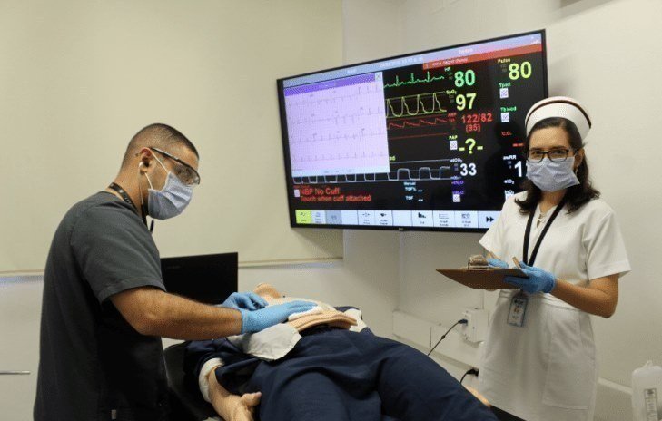 Simulación médica con monitor de signos vitales y dos profesionales de salud.