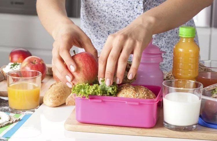 Mujer preparando una lonchera saludable en una mesa con alimentos como verduras, frutas y bebidas