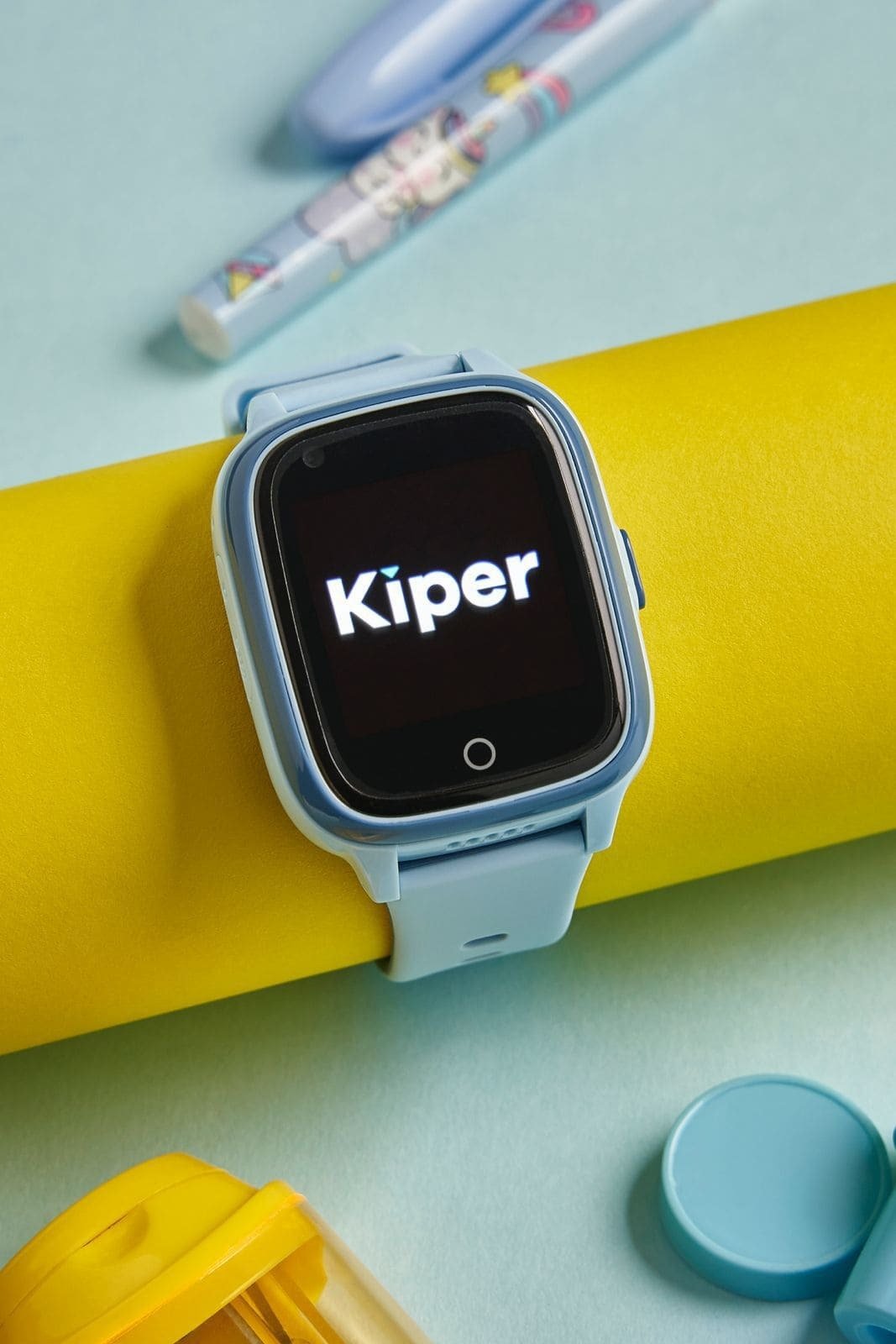 Un reloj inteligente para niños de la marca “Kiper”. El reloj tiene un diseño colorido, con correa azul clara y una pantalla negra donde se ve el logo. Está apoyado sobre un cilindro amarillo, rodeado de objetos infantiles como un marcador decorado y tapa