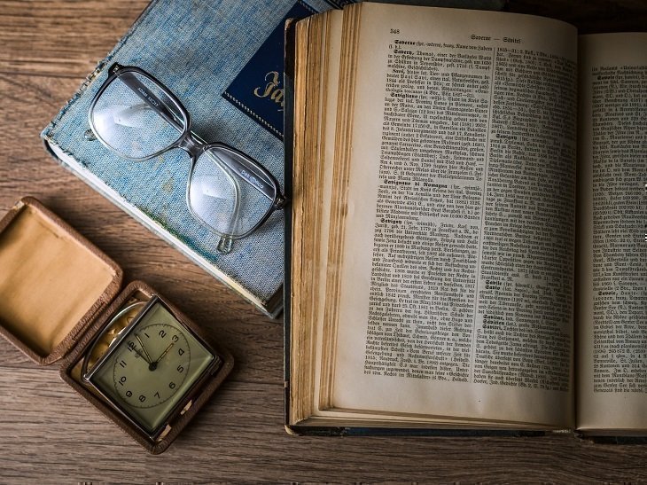 Sobre una superficie de madera se encuentran: un reloj, un cuaderno, un libro y unas gafas.