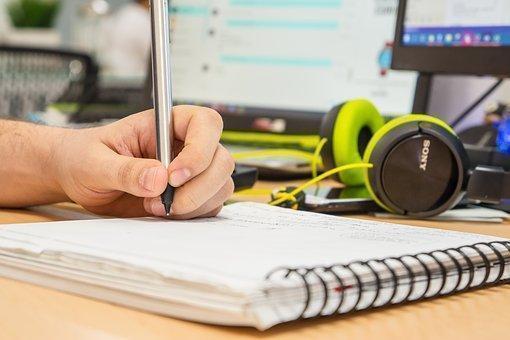 Sobre una mesa se encuentran unos audífonos verdes, dos pantallas de computador y una mano izquierda escribiendo sobre un cuaderno. 