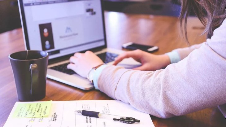 Mujer escribiendo en un computador portátil en un escritorio, al lado una taza de café y documentos