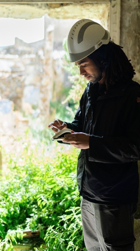 Un hombre con casco de obra mirando y usando una tableta mientras está junto a una pared, con vegetación al fondo.